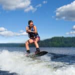 Young pretty slim brunette woman riding wakeboard on wave of motorboat in summer lake